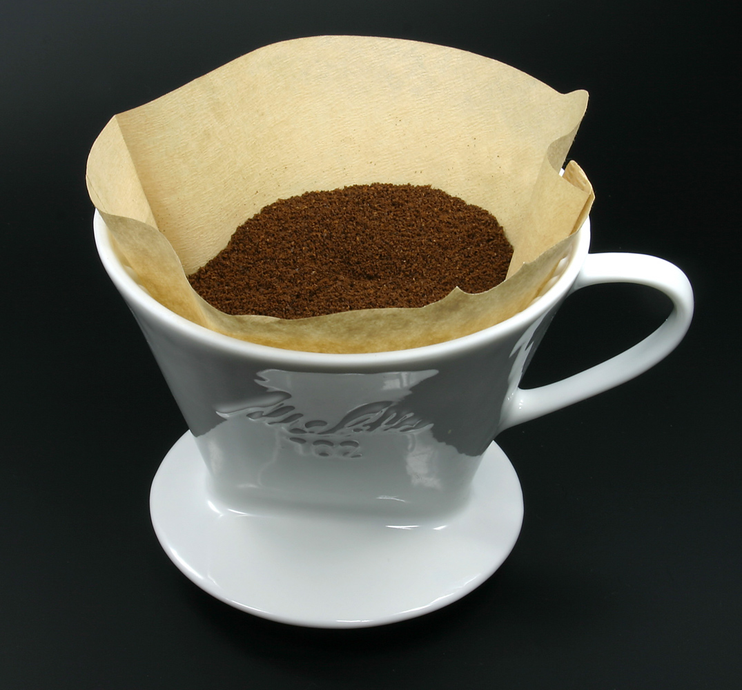 Portafiltro Melitta in porcellana con filtro di carta inserito e letto di caffè macinato pronto per l’estrazione pour-over, primo piano su fondo scuro.
